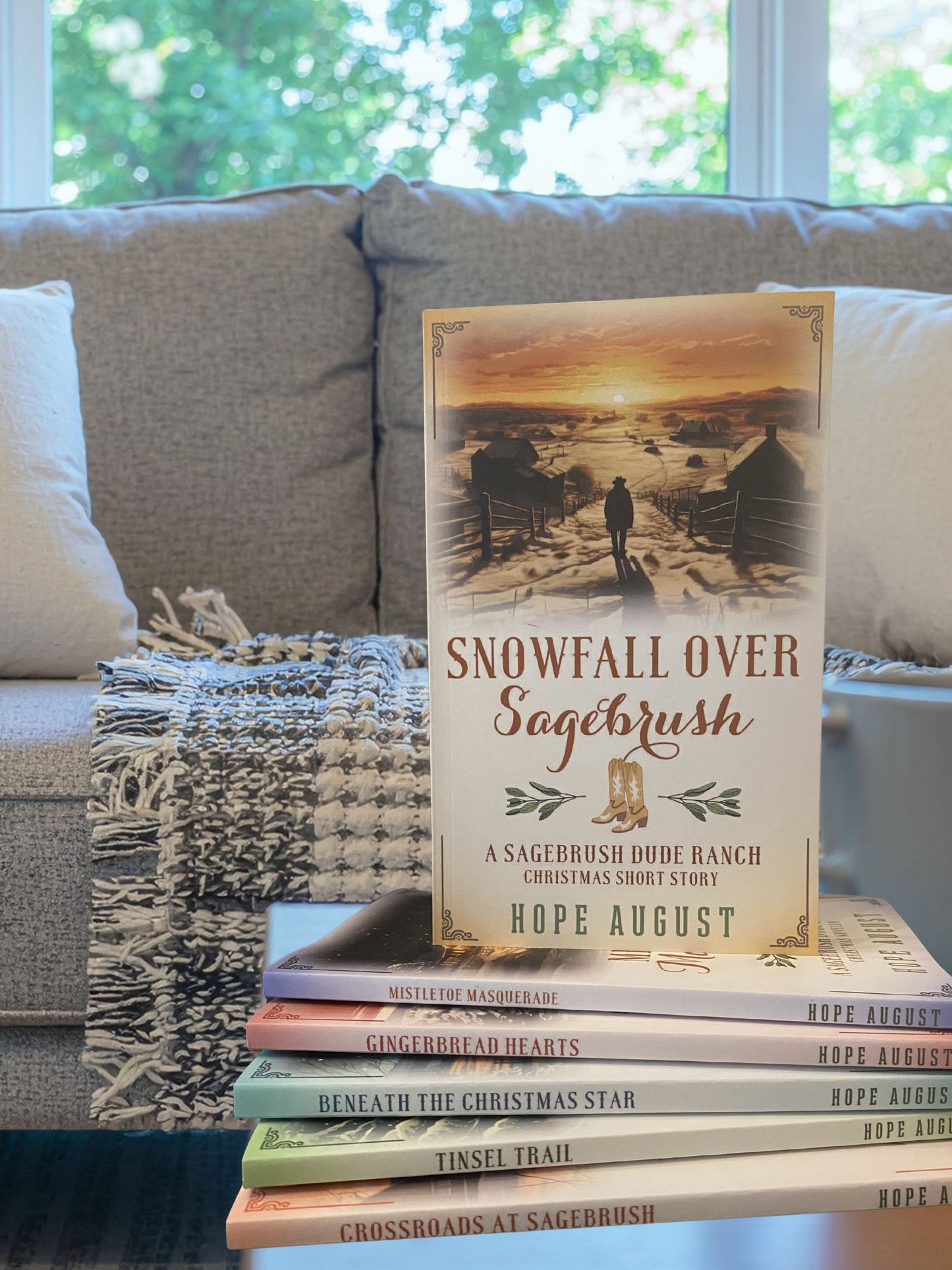Stack of books on a couch with 'Snowfall Over Sagebrush' by Hope August visible
